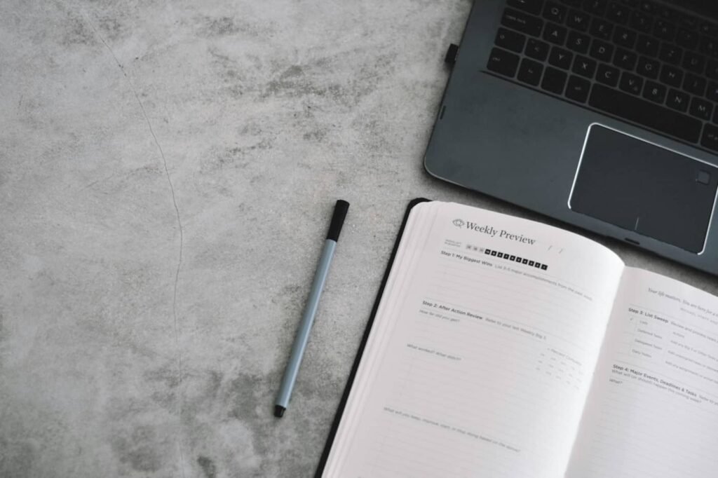 Otwarty planer tygodniowy obok laptopa na biurku – ustalanie priorytetów na dzień pracy w biurze.