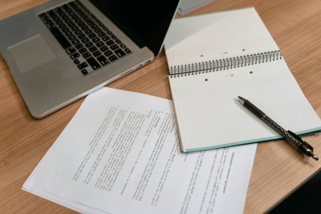 Otwarty notes z piórem, laptop i wydrukowane dokumenty tekstowe przygotowane do rocznej oceny pracownika.